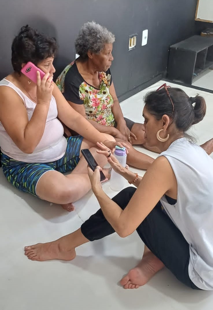 A imagem mostra três mulheres sentadas no chão, sendo que uma delas orienta as demais sobre o uso do aplicativo. 