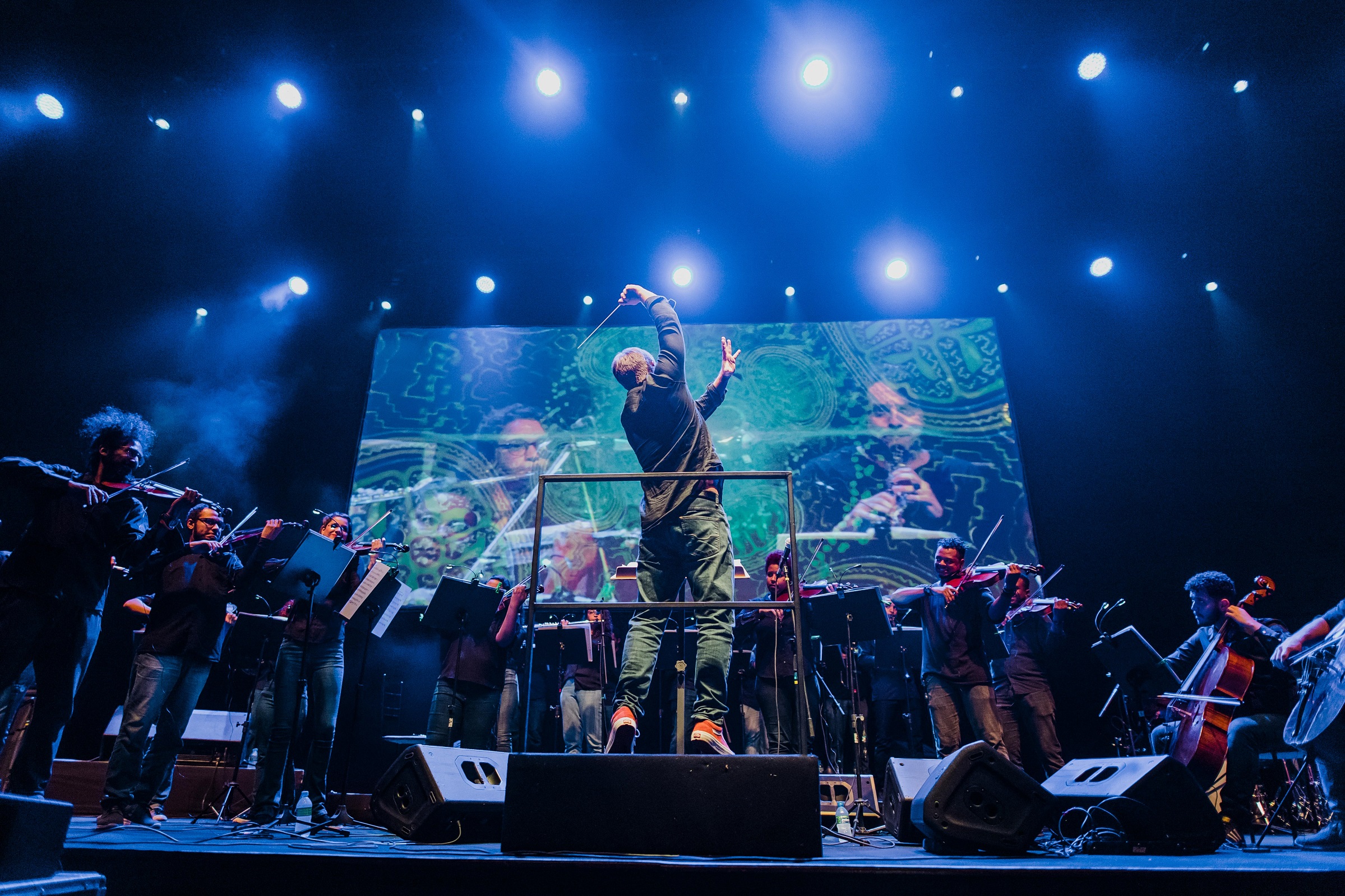 Foto de uma apresentação da orquestra onde aparecem o maestro e os músicos em um ambiente com luz azul