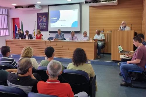 Foto exibe pessoas de costas, sentadas no auditório, e à frente autoridades sentadas à mesa, à esquerda, e o reitor da Ufes falando do púlpito, à direita