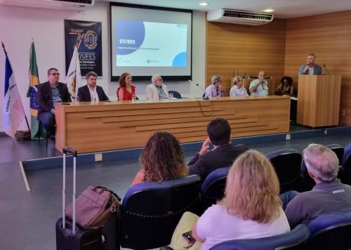 Foto exibe pessoas de costas, sentadas no auditório, e à frente autoridades sentadas à mesa, à esquerda, e o vice-reitor da Ufes falando do púlpito, à direita