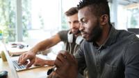 Foto de dois homens trabalhando diante de um notebook. Em primeiro plano, um homem negro. Em segundo plano, um homem branco.