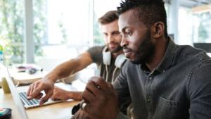 Foto de dois homens trabalhando diante de um notebook. Em primeiro plano, um homem negro. Em segundo plano, um homem branco.