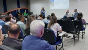 Foto das pessoas presentes no evento, de costas, sentadas nas cadeiras do auditório, com o reitor da Ufes à frente, sentado e falando ao microfone