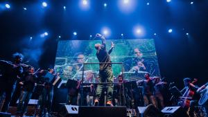 Foto de uma apresentação da orquestra onde aparecem o maestro e os músicos em um ambiente com luz azul