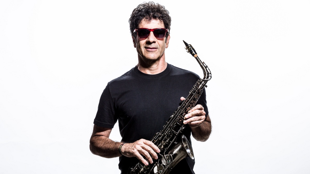 Foto do saxofonista George Israel. Ele veste camiseta preta, um óculos de aros avermelhados e segura o sax.