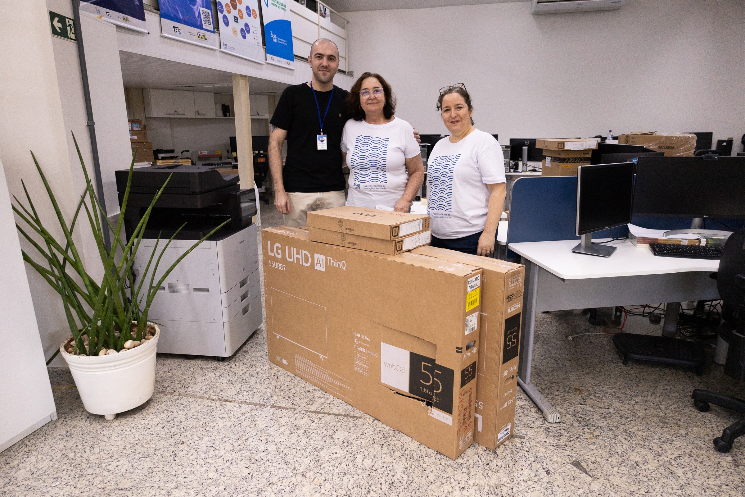 O coordenador do Polo de Baixo Guandú, Marcio Guilherme Marques, a superintendente de Educação a Distância, Maria Auxiliadora Corassa, e a diretora Acadêmica, Andreia Lins, posam com as caixas dos equipamentos na sala da Sead.