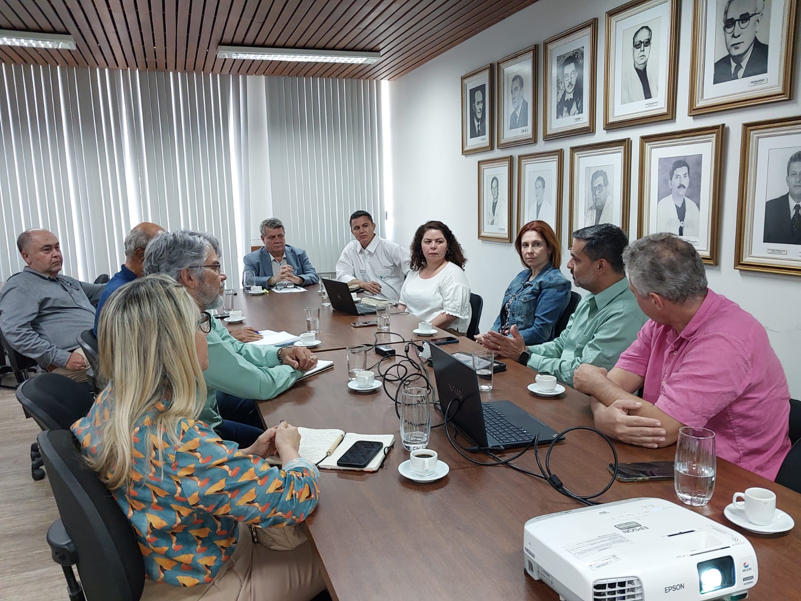 Foto da reunião onde os participantes estão sentados em volta de uma grande mesa de madeira. Em uma parede da sala, a galeria de fotos de ex-reitores da Ufes 