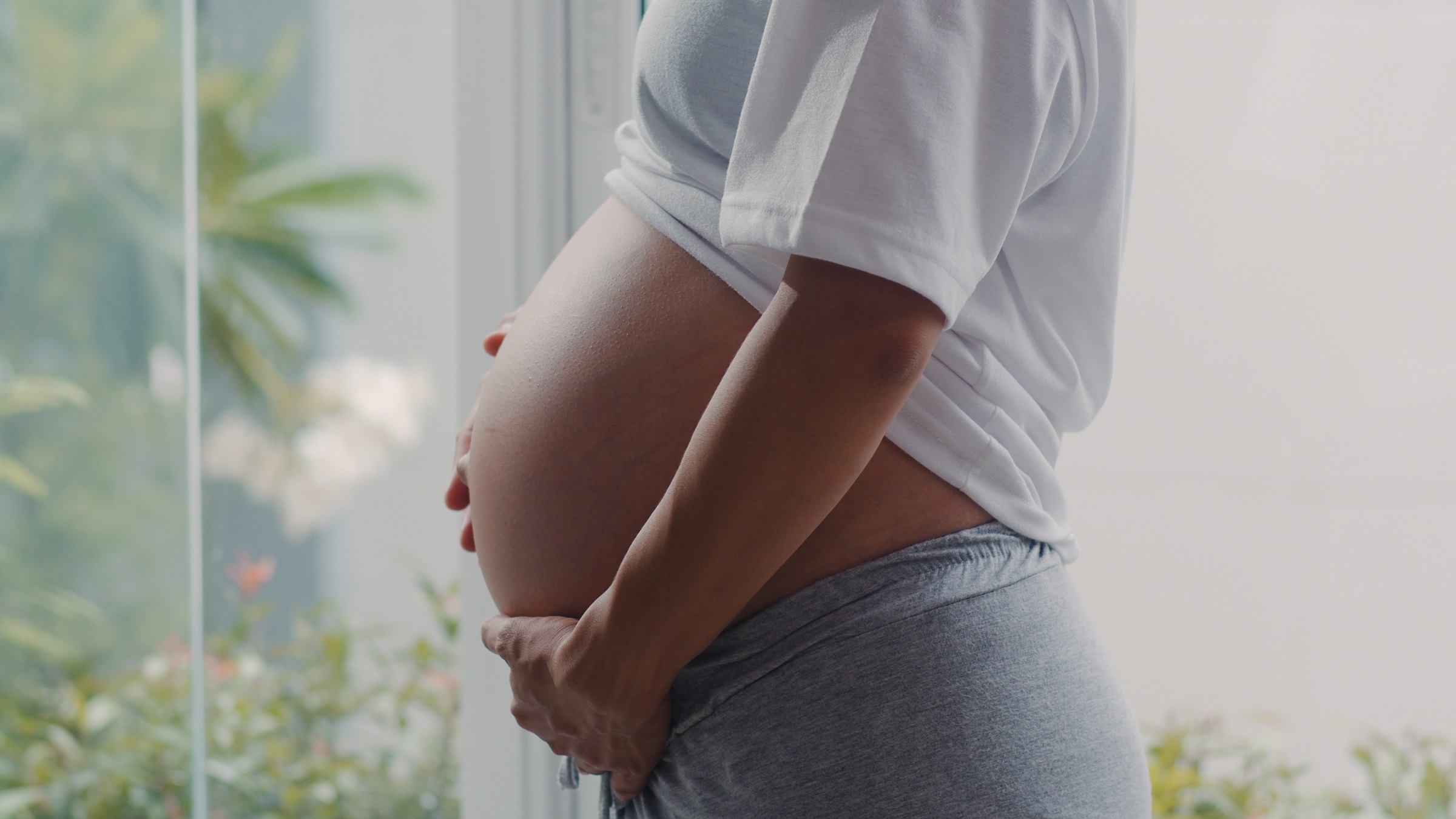 Perfil de uma mulher grávida com as mãos segurando a barriga, em um quarto claro