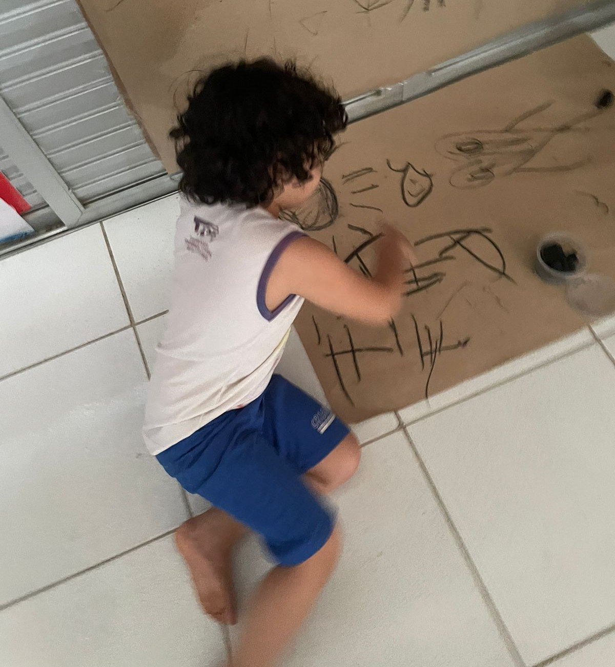 Foto de criança com uniforme da escola sentada no chão, desenhando sobre uma folha de papel pardo