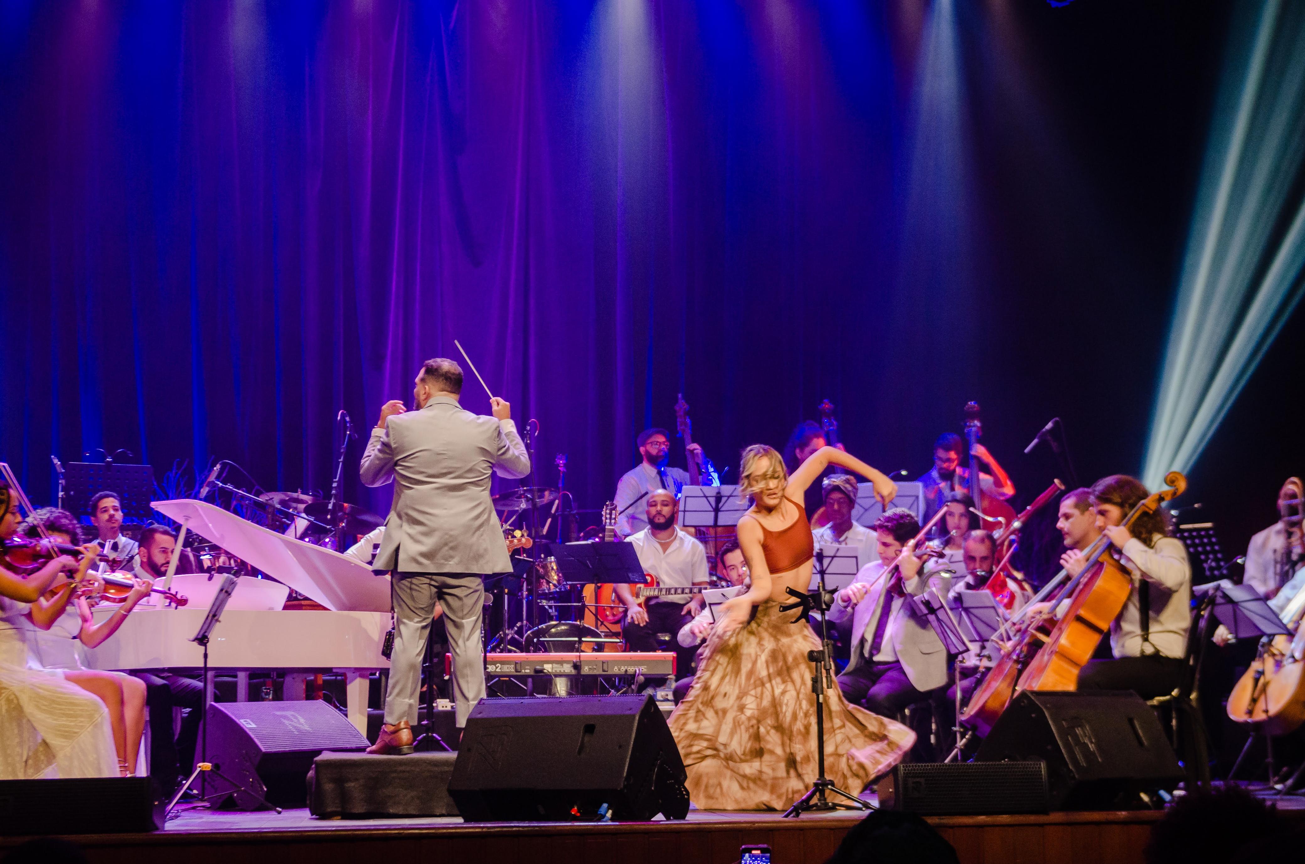 Foto da Orquestra se apresentando em um palco com uma mulher à frente, apresentando uma coreografia