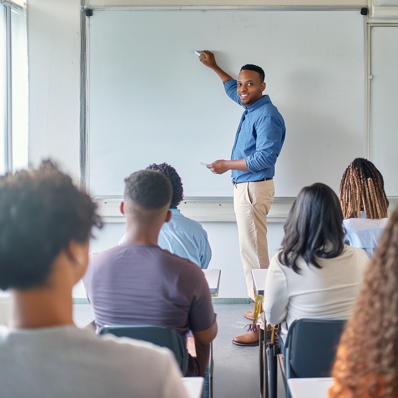 Professor em uma sala de aula escrevendo no quadro branco, de frente para a turma de jovens estudantes