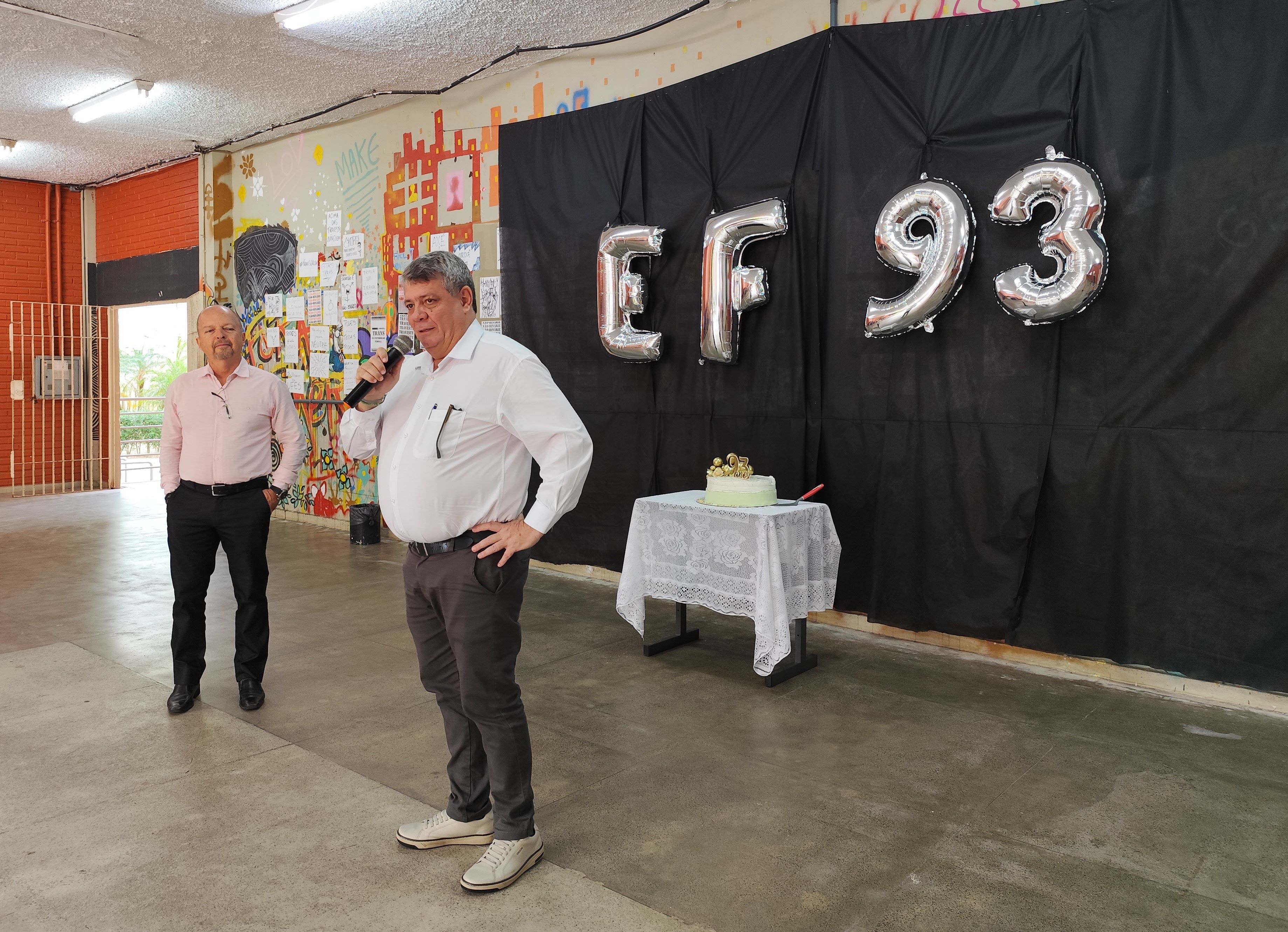 Foto do reitor e do diretor do Centro no hall. Ao fundo, um bolo e balões com as letras EF (de educação física) e o número  93