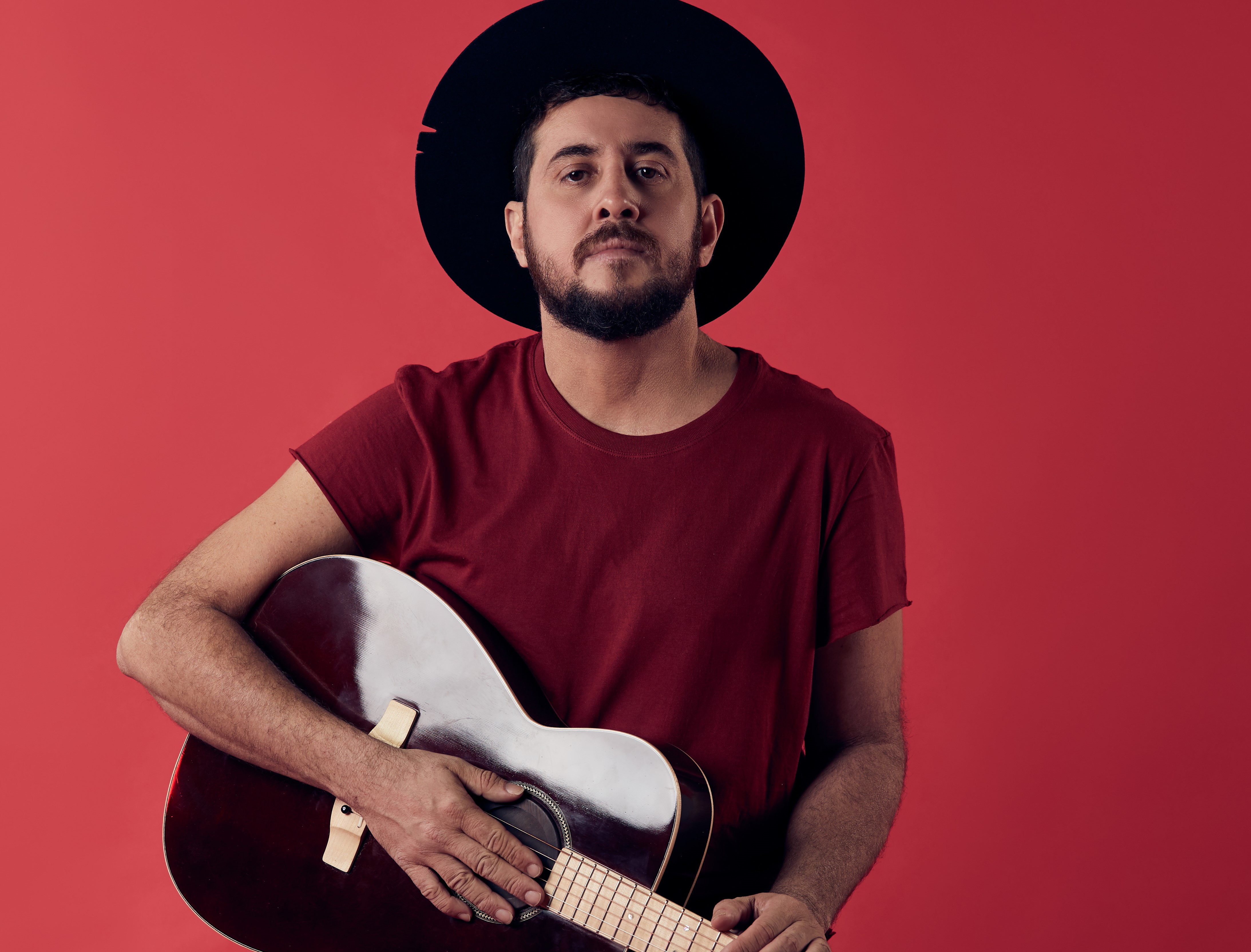 Foto do músico Rodrigo Suricato usando um chapéu preto e segurando um violão 