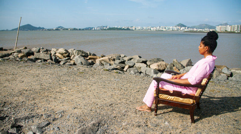 Foto da artista Rubiane Maia sentada em uma poltrona em um píer de frente para o mar, em uma cena do filme Presença