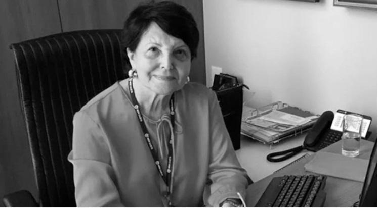 Foto de Maria Alice Lindenberg sentada em sua mesa de trabalho, onde é possível ver um teclado de computador, um telefone e um copo d´água