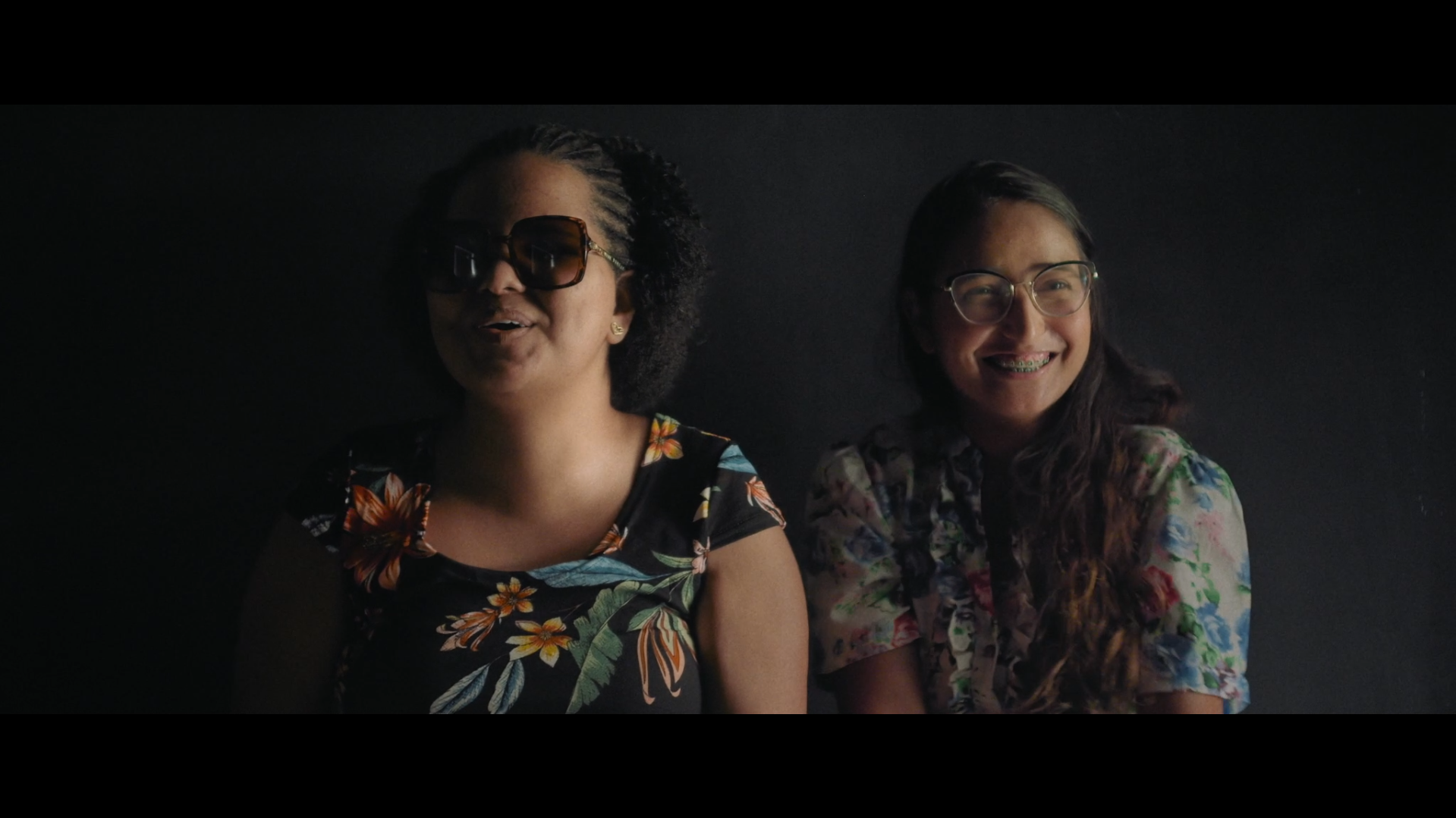 À esquerda, Cinthya de Oliveira em cena do filme Mão com Mão Cena do filme Mão com Mão, onde aparecem duas mulheres de frente para a câmera. Uma delas é Cinthya de Oliveira