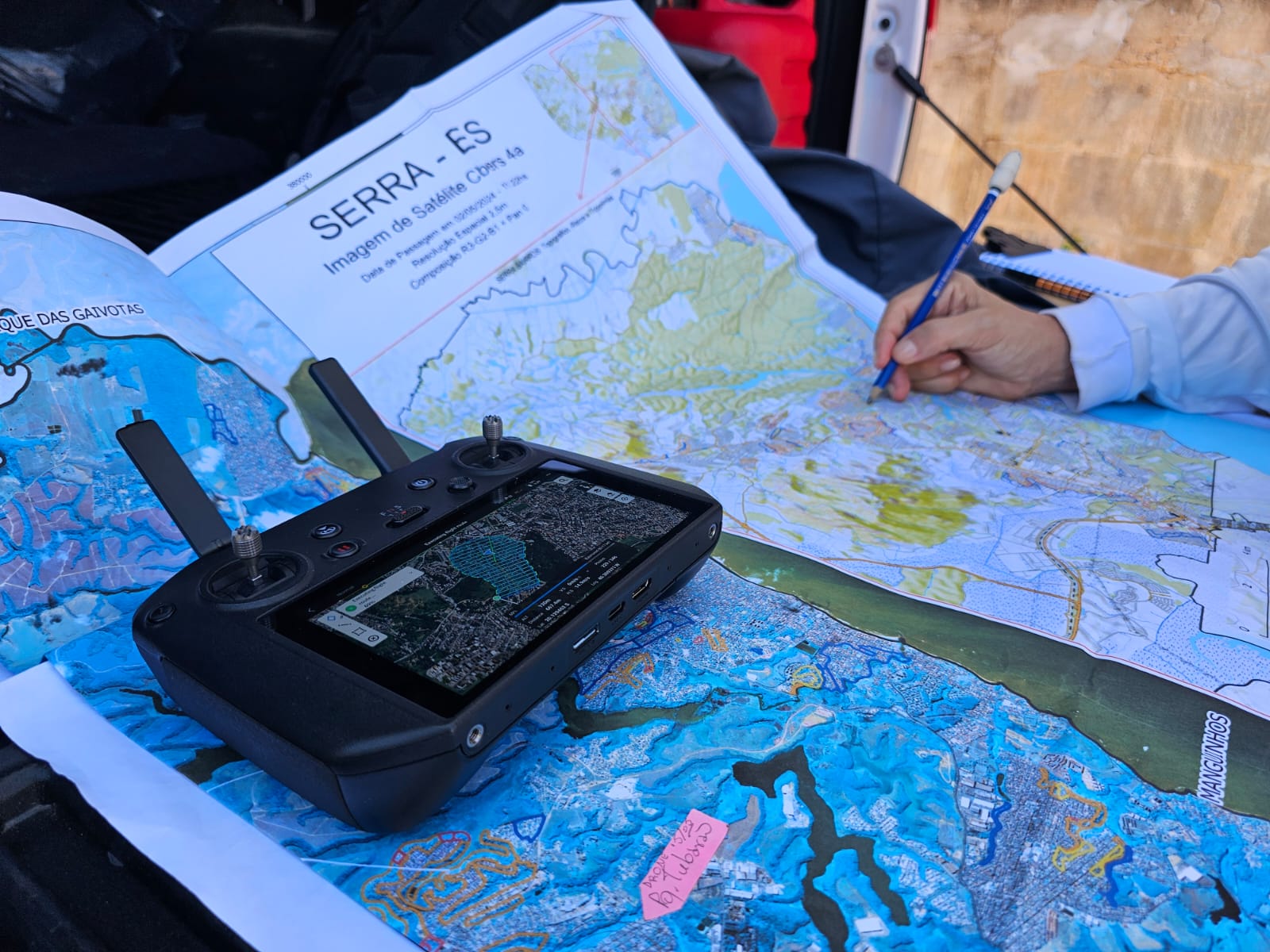 Foto de um mapa aberto do município da Serra com um controle remoto em cima e a mão de uma pessoa fazendo uma anotação a lápis sobre o mapa