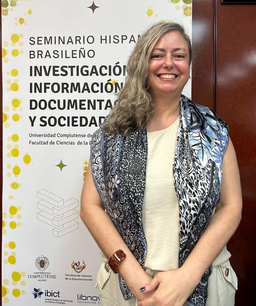 Foto da professora em pé, em frente a um banner de evento