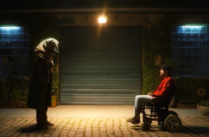 Cena de Memórias de um Esclerosado Cena de Memórias de um Esclerosado, na qual um homem em cadeira de rodas está de frente para uma pessoa em pé com uma cabeça de sapo