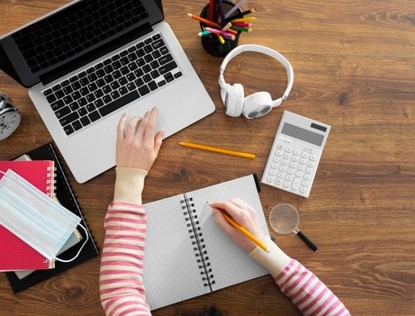 Imagem de uma pessoa estudante no computador. A foto, tirada do alto, mostra apenas os braços, o notebook, uma calculadora e o fone de ouvido