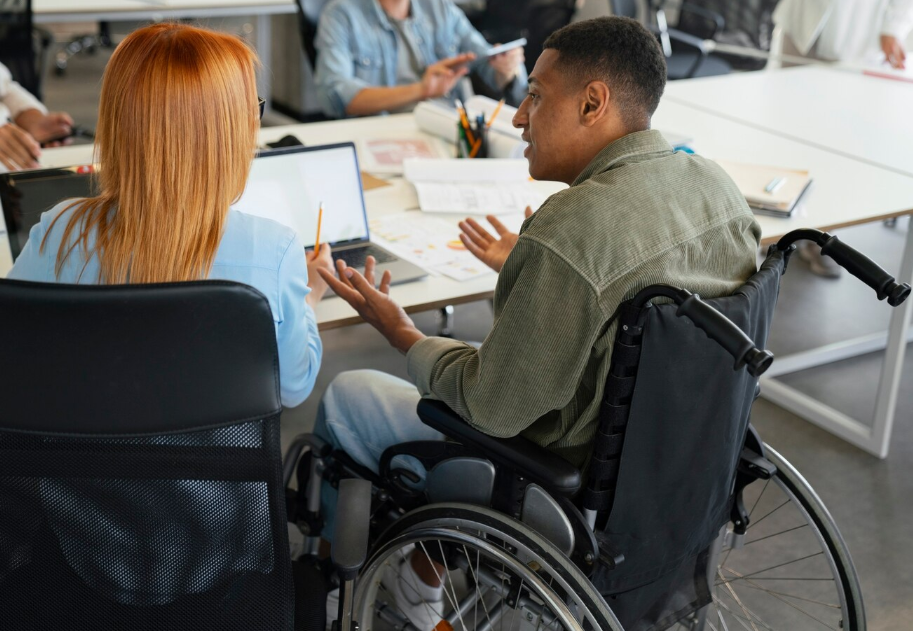 Foto de estudantes jovens conversando sentados à mesa, um deles está em uma cadeira de rodas