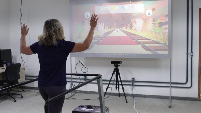 Esteira inteligente desenvolvida na Ufes Foto de uma mulher andando na esteira, tendo à frente um telão com a imagem projetada de um caminho