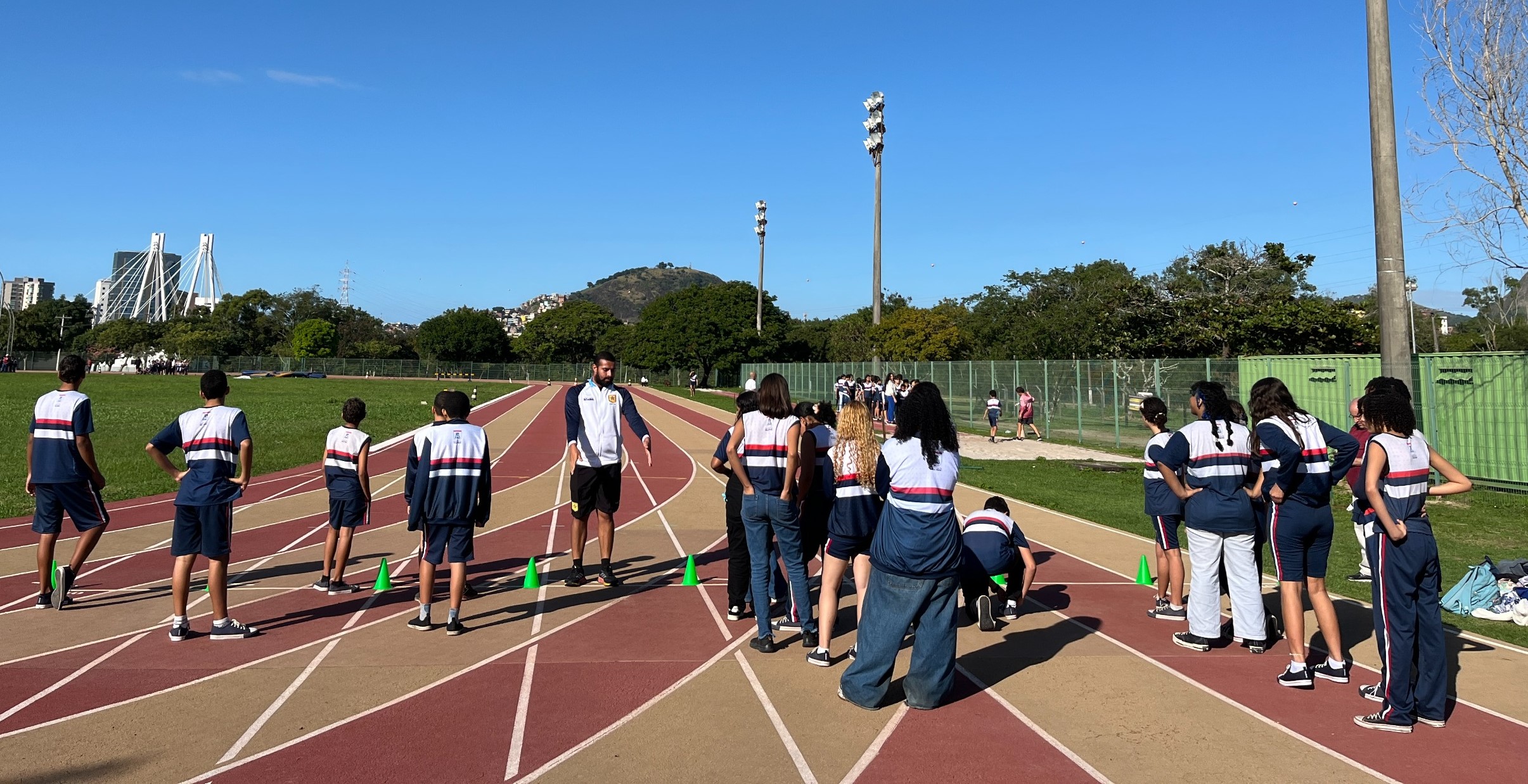 Alunos da rede municipal de Vila Velha participam do projeto Escola na Pista Foto dos alunos em pé na pista de atletismo, ouvido a explicação de um professor