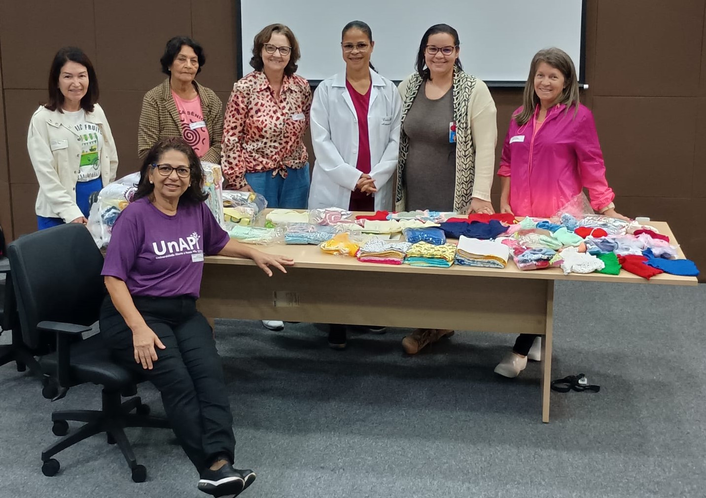 Foto da equipe do hospital com as crocheteiras, em volta de uma mesa onde estão as peças doadas