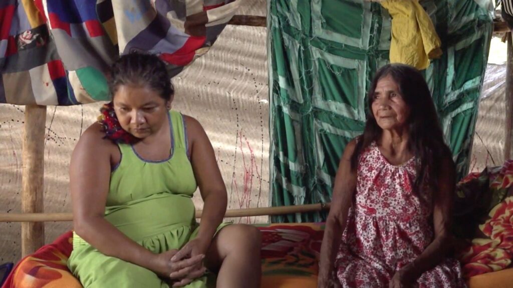 Cena de Yõg Ãtak: Meu Pai, Kaiowá Cena de Yõg Ãtak: Meu Pai, Kaiowá na qual aparecem duas mulheres indígenas sentadas no interior do que parece ser uma oca