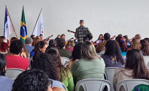 Foto da abertura do evento, com os servidores assistindo à apresentação da Orquestra de Violões, ao fundo