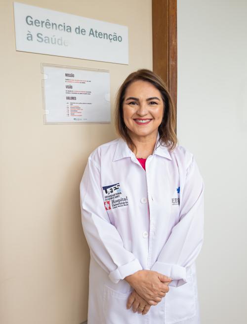 Foto da médica Valéria Valim vestindo um jaleco com a marca da Ufes e do Hospital Universitário, em pé, em frente à porta da Gerência de Atenção à Saúde