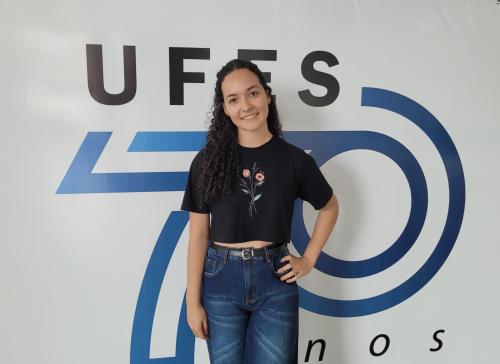 Foto da estudante Gabrielly dos Santos em pé, em frente à logomarca de comemoração dos 70 anos da Ufes
