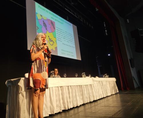 Foto da professora da Universidade do Estado da Bahia Marcea Sales, em pé, no palco do Teatro, ministrando a aula inaugural. 