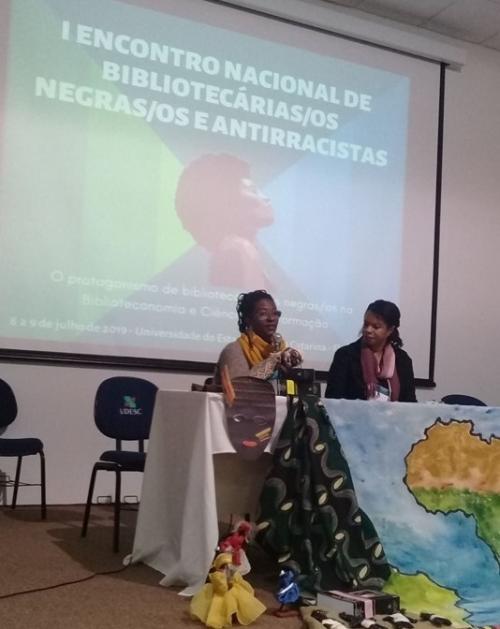 Foto da professora compondo a mesa principal de um evento, falando ao microfone 