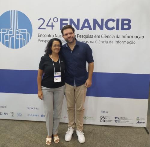 Foto de dois autores do artigo, Daniela Lemos e Dirceu Macedo, em frente a um banner do evento