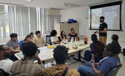 Foto dos estudantes reunidos em torno da mesa, olhando para a apresentação projetada na parece. Em pé, o instrutor fala para os estudantes.
