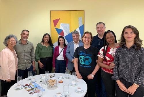 Os participantes da reunião em pé, posando para a foto em volta de uma mesa redonda