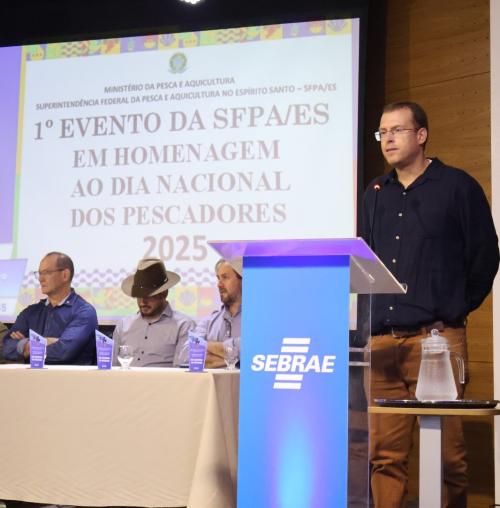 O professor Joelson Musiello no púlpito, ao lado da mesa de abertura do evento onde estão outros três homens.