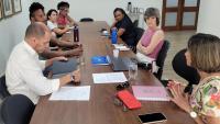 Foto da reunião, onde todos os participantes estão em uma sala de paredes brancas, sentados em torno de uma grande mesa de madeira 