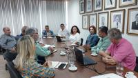 Foto da reunião onde os participantes estão sentados em volta de uma grande mesa de madeira. Em uma parede da sala, a galeria de fotos de ex-reitores da Ufes 