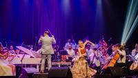 Foto da Orquestra se apresentando em um palco com uma mulher à frente, apresentando uma coreografia