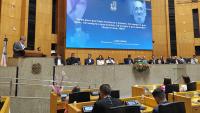 Foto do reitor no púlpito da Assembleia Legislativa do Espírito Santo. Ao fundo, a mesa principal com autoridades e, ao fundo, um telão no qual está projetada a figura de Paulo Freire.