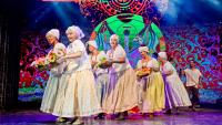 Foto de mulheres do Grupo Andora em uma apresentação no palco. Elas vestem blusas brancas, saias rodadas com estampa florida em tom amarelo claro. Nas mãos carregam flores como oferendas..