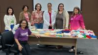 Foto da equipe do hospital com as crocheteiras, em volta de uma mesa onde estão as peças doadas