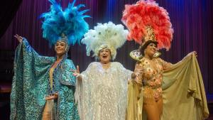 Imagem de três atrizes vestidas de vedetes, com roupas brilhosas e plumas na cabeça, no palco de um teatro