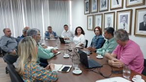 Foto da reunião onde os participantes estão sentados em volta de uma grande mesa de madeira. Em uma parede da sala, a galeria de fotos de ex-reitores da Ufes