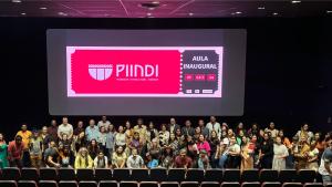 Abertura da aula inaugural do PIINDI Foto da abertura da aula inaugural, com todos os presentes posando para a foto. Atrás, projetada na tela do cinema, a logomarca do curso e a data da aula.