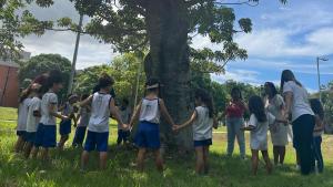 Uma das atividades do Núcleo realizadas com alunos do Colégio de Aplicação Criarte Foto de crianças de mãos dadas em volta de uma árvore, em uma das atividade do Núcleo