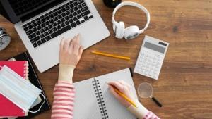 Imagem de uma pessoa estudante no computador. A foto, tirada do alto, mostra apenas os braços, o notebook, uma calculadora e o fone de ouvido