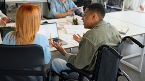 Foto de estudantes jovens conversando sentados à mesa, um deles está em uma cadeira de rodas